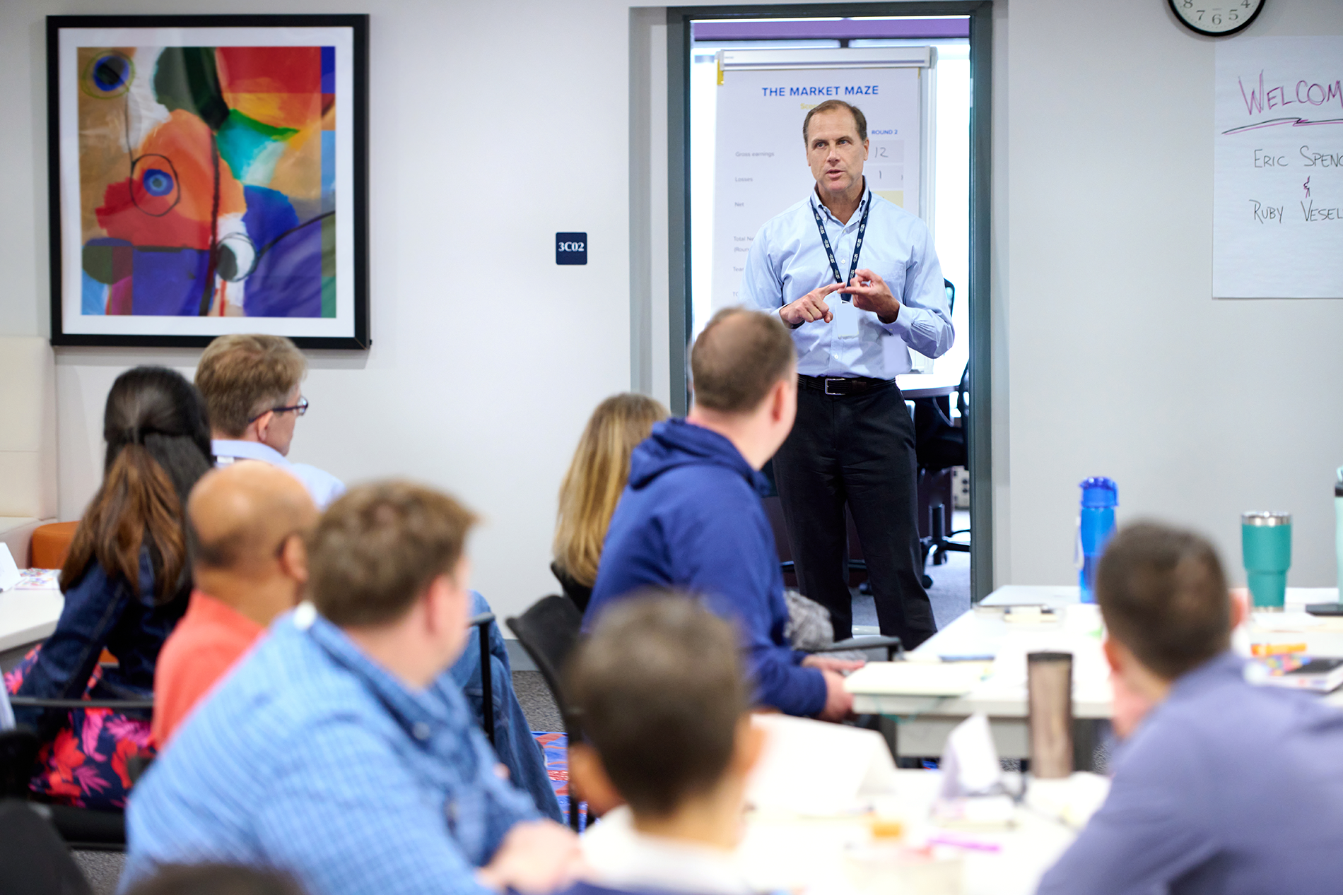A facilitator engages a group during a workshop, discussing strategies with participants seated at tables.
