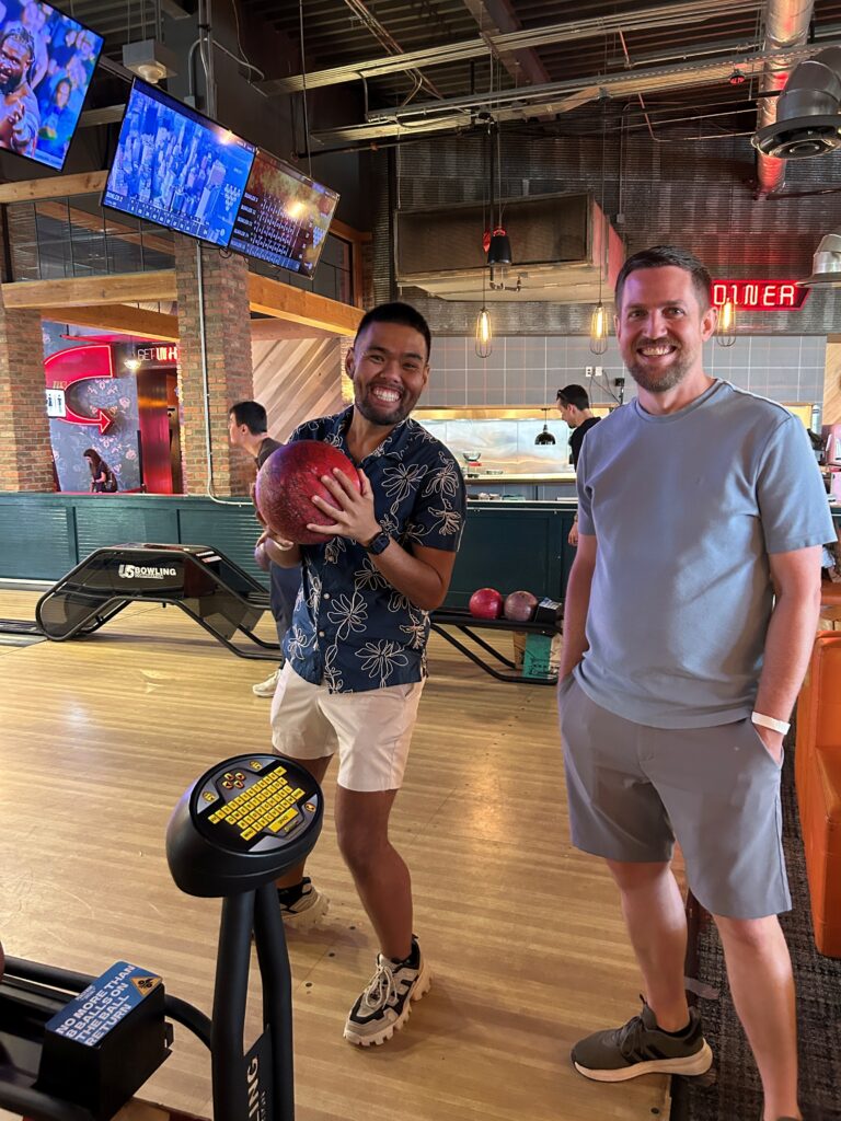 Two friends smile while bowling, one holding a bowling ball, in a lively bowling alley setting with screens above.